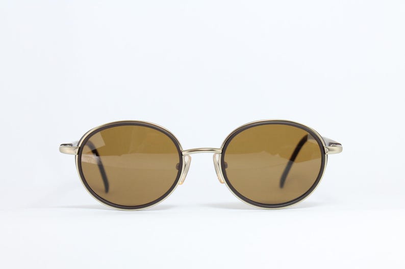 K&ouml;nnte beinhalten: Runde Sonnenbrille mit braunen Gl&auml;sern und einem goldfarbenen Metallrahmen. Die Gl&auml;ser sind von einem dunkelbraunen Rand umgeben. Die B&uuml;gel sind d&uuml;nn und goldfarben. Die Sonnenbrille ist vor einem wei&szlig;en Hintergrund platziert.
