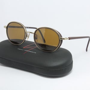 K&ouml;nnte beinhalten: Runde Sonnenbrille mit braunen Gl&auml;sern und goldenem Rahmen liegt auf einem schwarzen Etui. Die B&uuml;gel sind braun. Auf dem Etui steht der Text "YOHJI YAMAMOTO" in Wei&szlig; und Rot.