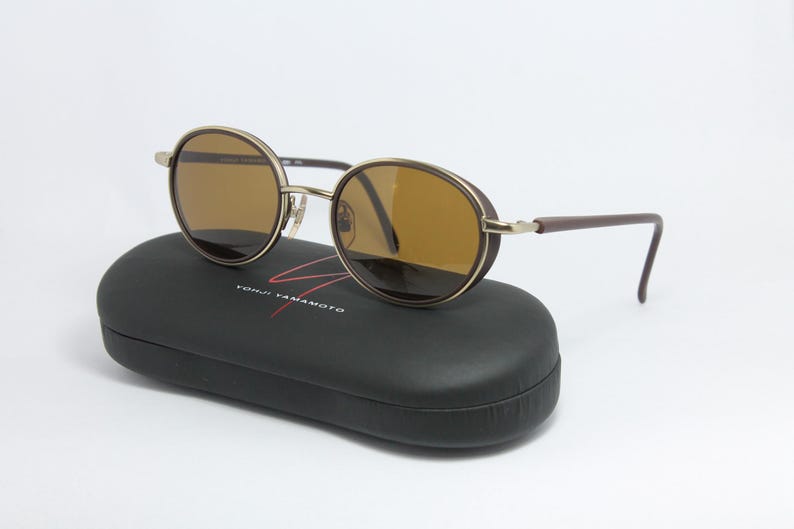 K&ouml;nnte beinhalten: Runde Sonnenbrille mit braunen Gl&auml;sern und goldenem Rahmen liegt auf einem schwarzen Etui. Die B&uuml;gel sind braun. Auf dem Etui steht der Text "YOHJI YAMAMOTO" in Wei&szlig; und Rot.