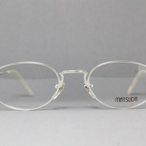 May include: Silver-framed eyeglasses with oval lenses. The frames have a delicate, minimalist design with the brand name "MATSUDA" printed on the right lens. The temples are a light beige color.