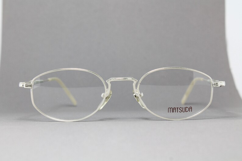 May include: Silver-framed eyeglasses with oval lenses. The frames have a delicate, minimalist design with the brand name "MATSUDA" printed on the right lens. The temples are a light beige color.