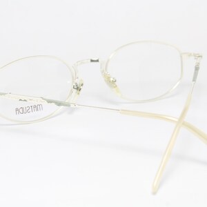 May include: A pair of vintage Matsuda eyeglasses with a light-colored metal frame and oval lenses. The temples are a light beige color. The brand name "MATSUDA" is visible on the frame. The glasses are set against a white background.
