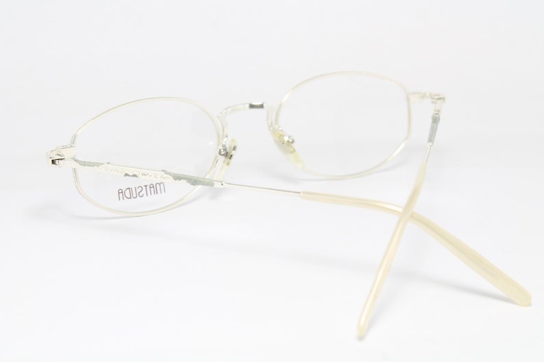 May include: A pair of vintage Matsuda eyeglasses with a light-colored metal frame and oval lenses. The temples are a light beige color. The brand name "MATSUDA" is visible on the frame. The glasses are set against a white background.