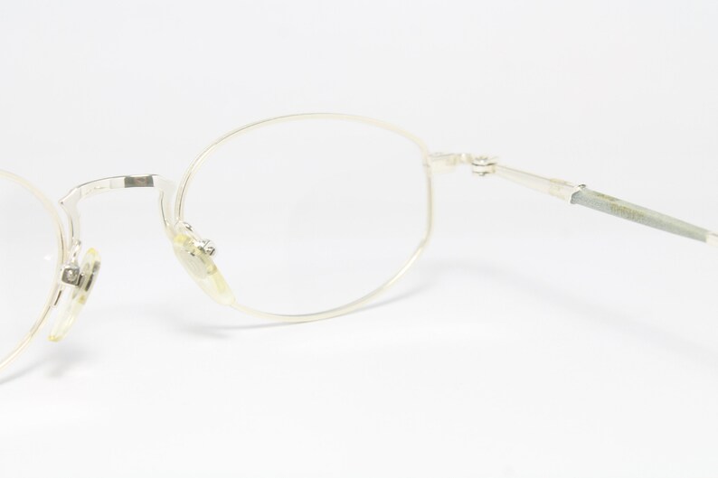 May include: Close-up of a pair of silver-rimmed eyeglasses with clear lenses. The glasses have a classic round shape and are set against a white background. The frames are thin and delicate.