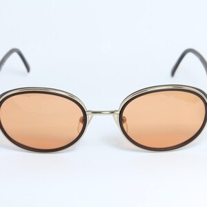 May include: Oval-shaped sunglasses with orange-tinted lenses. The frames are a combination of silver-tone metal, dark brown accents, and black temples. The sunglasses have a classic, retro design.