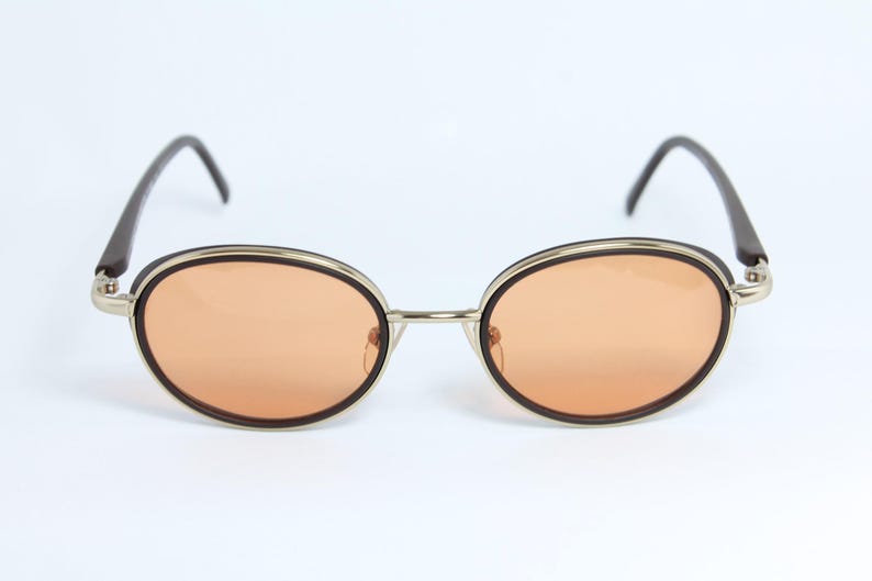 May include: Oval-shaped sunglasses with orange-tinted lenses. The frames are a combination of silver-tone metal, dark brown accents, and black temples. The sunglasses have a classic, retro design.