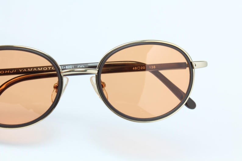 May include: Round sunglasses with a gold frame and brown temples. The lenses are a light orange color. The text "YOHJI YAMAMOTO" is visible on the frame.