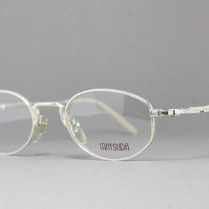 May include: Silver-toned Matsuda eyeglasses with oval lenses. The frames have a delicate, minimalist design with the brand name printed on the right lens. The temples feature an ornate, textured pattern.
