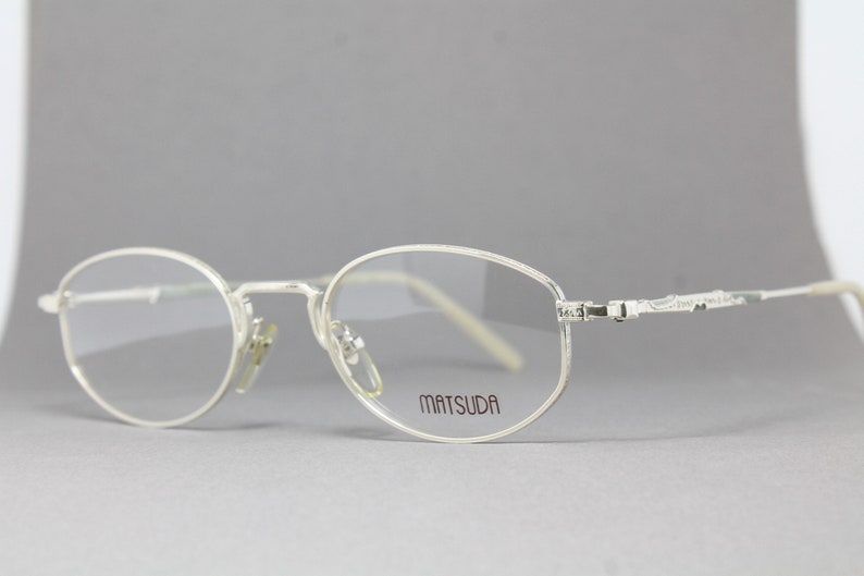 May include: Silver-toned Matsuda eyeglasses with oval lenses. The frames have a delicate, minimalist design with the brand name printed on the right lens. The temples feature an ornate, textured pattern.