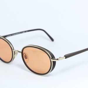 May include: Oval-shaped sunglasses with orange-tinted lenses. The frames are a combination of silver, black, and brown. The arms are dark brown, and the lenses are a warm orange color. The glasses have a retro, vintage style.