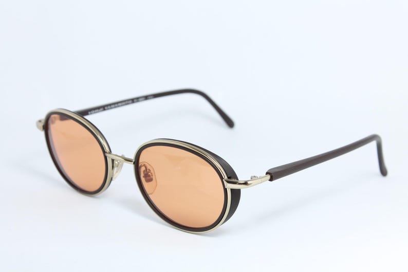 May include: Oval-shaped sunglasses with orange-tinted lenses. The frames are a combination of silver, black, and brown. The arms are dark brown, and the lenses are a warm orange color. The glasses have a retro, vintage style.
