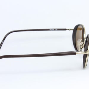K&ouml;nnte beinhalten: Braune ovale Sonnenbrille mit goldenem Rahmen und braunen B&uuml;geln. Die Gl&auml;ser sind hellbraun. Die Brille ist vor einem wei&szlig;en Hintergrund platziert.