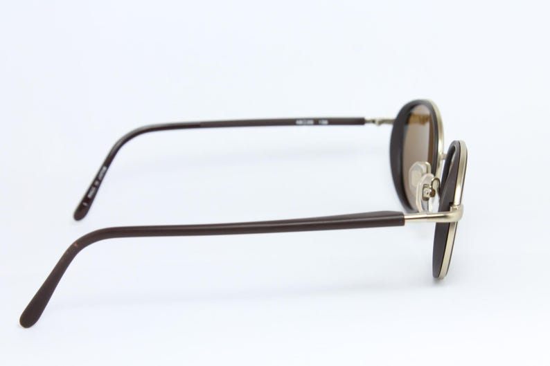 K&ouml;nnte beinhalten: Braune ovale Sonnenbrille mit goldenem Rahmen und braunen B&uuml;geln. Die Gl&auml;ser sind hellbraun. Die Brille ist vor einem wei&szlig;en Hintergrund platziert.