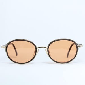May include: Oval-shaped sunglasses with a silver frame and brown rims. The lenses are a light orange color. The arms are dark brown. The sunglasses are set against a white background.