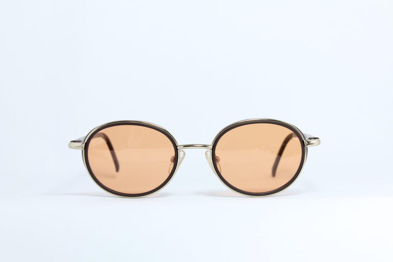 May include: Oval-shaped sunglasses with a silver frame and brown rims. The lenses are a light orange color. The arms are dark brown. The sunglasses are set against a white background.