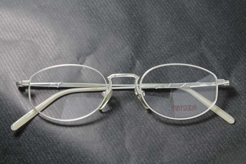 May include: Silver-toned eyeglasses with oval lenses and a bridge. The temples and frame are thin and metallic. The brand name "MATSUDA" is printed on the right lens.