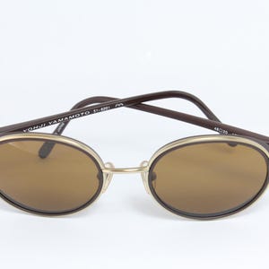 K&ouml;nnte beinhalten: Ovale Sonnenbrille mit braunen Gl&auml;sern und einem goldfarbenen Rahmen. Die dunkelbraunen B&uuml;gel tragen den Schriftzug "YOHJI YAMAMOTO 51-6201". Klassisches, Vintage-Design.