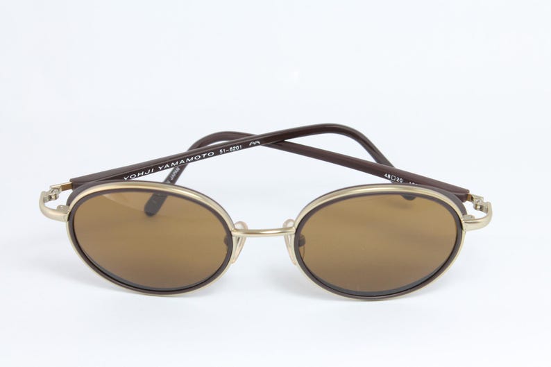 K&ouml;nnte beinhalten: Ovale Sonnenbrille mit braunen Gl&auml;sern und einem goldfarbenen Rahmen. Die dunkelbraunen B&uuml;gel tragen den Schriftzug "YOHJI YAMAMOTO 51-6201". Klassisches, Vintage-Design.