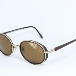 K&ouml;nnte beinhalten: Ovale Sonnenbrille mit braunen Gl&auml;sern und einem silbernen Rahmen. Die R&auml;nder sind schwarz und die B&uuml;gel dunkelbraun. Die Sonnenbrille ist vor einem wei&szlig;en Hintergrund platziert.