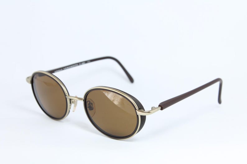 K&ouml;nnte beinhalten: Ovale Sonnenbrille mit braunen Gl&auml;sern und einem silbernen Rahmen. Die R&auml;nder sind schwarz und die B&uuml;gel dunkelbraun. Die Sonnenbrille ist vor einem wei&szlig;en Hintergrund platziert.