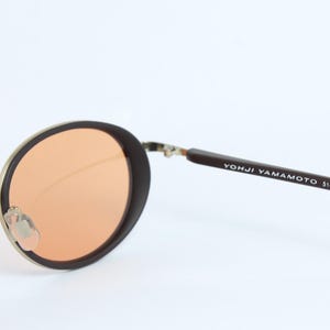May include: Round sunglasses with brown frames and orange lenses. The metal frame is gold-colored. The text "YOHJI YAMAMOTO 51-8201" is printed on the brown arm of the sunglasses.