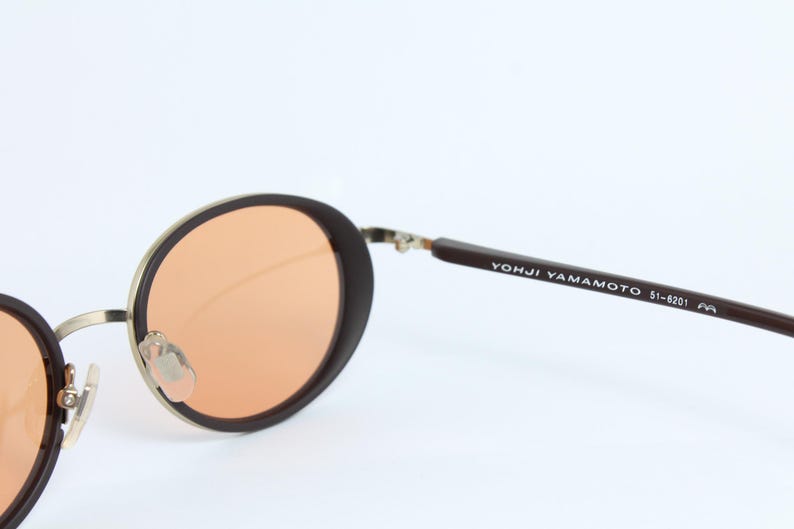 May include: Round sunglasses with brown frames and orange lenses. The metal frame is gold-colored. The text "YOHJI YAMAMOTO 51-8201" is printed on the brown arm of the sunglasses.