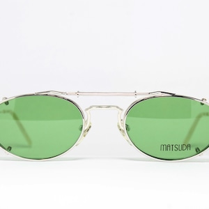 May include: Silver-framed sunglasses with green lenses. The glasses have a double bridge and a rectangular shape. The brand name "MATSUDA" is printed on the right lens. The sunglasses are set against a white background.