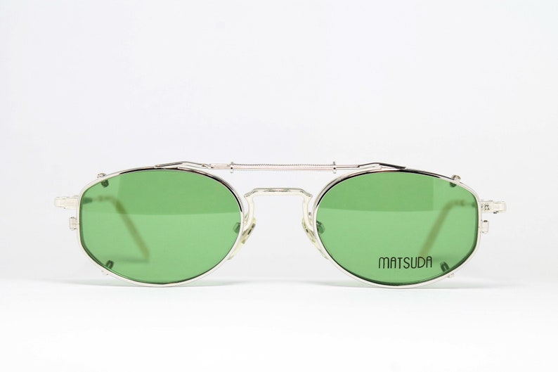 May include: Silver-framed sunglasses with green lenses. The glasses have a double bridge and a rectangular shape. The brand name "MATSUDA" is printed on the right lens. The sunglasses are set against a white background.