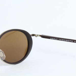 K&ouml;nnte beinhalten: Runde Sonnenbrille mit braunen Gl&auml;sern und einem dunkelbraunen Rahmen. Die Metallb&uuml;gel sind silberfarben. Der Text "YOHJI YAMAMOTO 51-6201" ist auf dem rechten B&uuml;gel aufgedruckt.