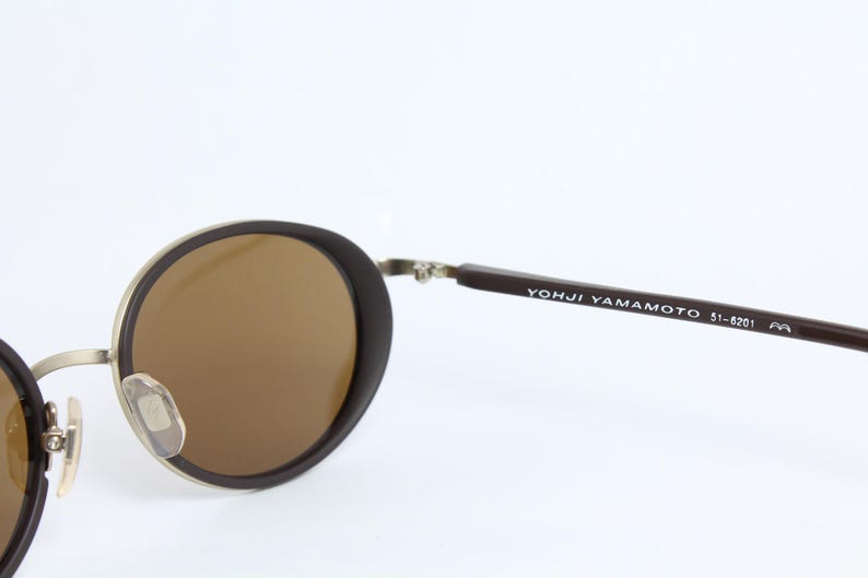 K&ouml;nnte beinhalten: Runde Sonnenbrille mit braunen Gl&auml;sern und einem dunkelbraunen Rahmen. Die Metallb&uuml;gel sind silberfarben. Der Text "YOHJI YAMAMOTO 51-6201" ist auf dem rechten B&uuml;gel aufgedruckt.