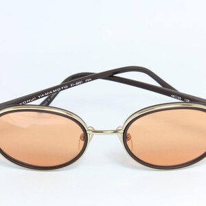 May include: Oval-shaped sunglasses with orange-tinted lenses. The frames are a combination of gold-tone metal and dark brown accents. The arms are dark brown with the text "YOHJI YAMAMOTO 51-8201" printed on the left arm. The lens width is 48mm.