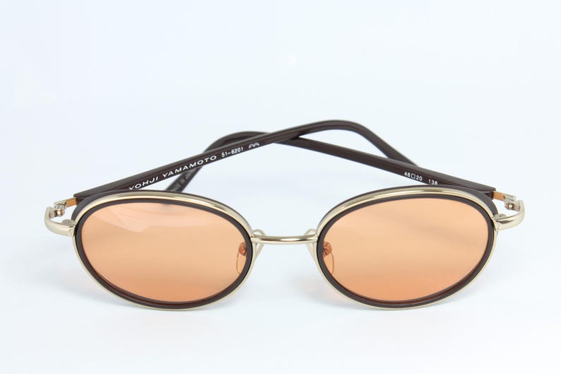 May include: Oval-shaped sunglasses with orange-tinted lenses. The frames are a combination of gold-tone metal and dark brown accents. The arms are dark brown with the text "YOHJI YAMAMOTO 51-8201" printed on the left arm. The lens width is 48mm.