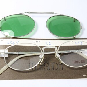 May include: Silver-framed Matsuda eyeglasses with a clip-on green lens attachment. The glasses have a classic oval shape and are displayed with their original packaging. The packaging includes the brand name and the text "Made in Japan".