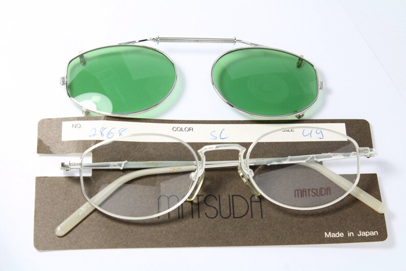 May include: Silver-framed Matsuda eyeglasses with a clip-on green lens attachment. The glasses have a classic oval shape and are displayed with their original packaging. The packaging includes the brand name and the text "Made in Japan".