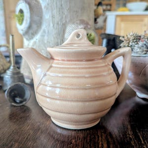 May include: A small, tan ceramic teapot with a lid and handle. The teapot has horizontal ridges and sits on a dark wooden surface. Other objects, including a lamp and a decorative pot, are in the background.