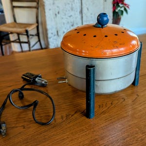 May include: A vintage orange and silver electric popcorn maker with a blue knob on top. The popcorn maker has four black legs and a cord with a plug.