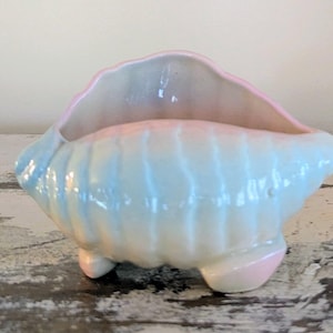 May include: A ceramic seashell-shaped planter in pastel colors. The planter has a light blue exterior with pink and yellow accents, and a pink rim. It rests on three small feet.