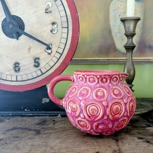 May include: A pink ceramic pitcher with a handle and a textured surface of concentric circles. The pitcher is set on a wooden surface, with a vintage clock and a candlestick in the background.