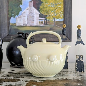 May include: A cream-colored ceramic teapot with a rounded body, a curved handle, and a spout. The teapot features two round, raised decorative elements and a lid with a small knob. The design includes vertical, wavy lines. A black jug is behind the teapot.