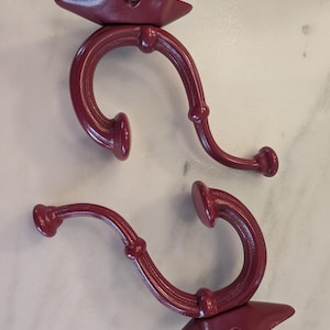 May include: Two burgundy-colored metal wall hooks. Each hook has a curved design with a decorative base and a small knob at the end. The hooks are designed for hanging items and add a touch of elegance to any space.