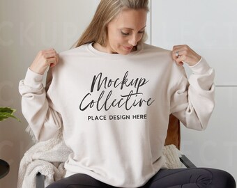 May include: A beige sweatshirt with the text "Mockup Collective" in black script. The sweatshirt is being held up by a person. The person is sitting in a chair. The background includes a white door and a small side table with a plant.