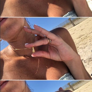 May include: A gold chain necklace with a small gold pendant shaped like a hand. The necklace is worn by a person with a tan complexion. The person is wearing a light blue tank top.