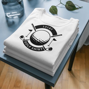 Puede incluir: Una camiseta blanca con un dise&ntilde;o gr&aacute;fico en blanco y negro de una bola de golf, palos de golf y el texto "Lucky Golf Shirt".
