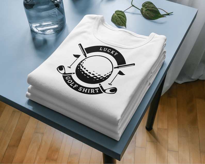 Puede incluir: Una camiseta blanca con un dise&ntilde;o gr&aacute;fico en blanco y negro de una bola de golf, palos de golf y el texto "Lucky Golf Shirt".