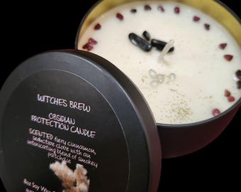 May include: A black candle tin with a gold interior, labeled "Witches Brew Obsidian Protection Candle." The candle wax is cream-colored, adorned with small red and black stones. The scent is described as fiery cinnamon, clove, and patchouli.