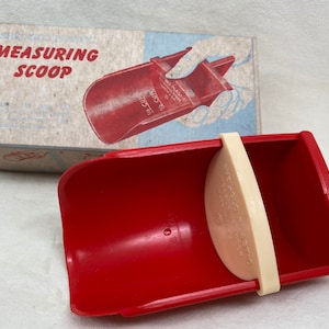 May include: A red plastic measuring scoop with a cream-colored insert, and a handle. The box behind it reads "Measuring Scoop" in red letters. The scoop is marked for 1/4 cup. Kitchen utensil.