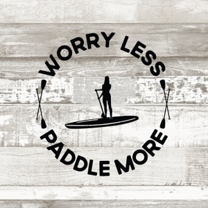 May include: Black and white graphic design with the text "Worry Less Paddle More" in a circle. A silhouette of a person paddleboarding is in the center of the circle. Two crossed paddles are on either side of the circle.