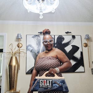 May include: A woman wearing a brown polka dot dress and holding a denim handbag. The handbag has a wooden handle and the word "GOAT" with "GOD OVER ALL THINGS" printed on it. She is also wearing jeweled glasses.