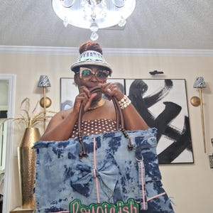 May include: A large denim tote bag with a bow and the word "Bougieish" in green and pink. The bag has brown leather handles and pink stitching. The denim has a blue tie-dye effect.