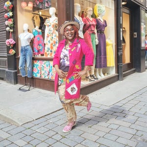 May include: A person wearing a bright pink coat with patches and the word "FASHION" on it, patterned pants, and pink shoes. The person is standing on a brick sidewalk in front of a store window displaying mannequins wearing dresses.